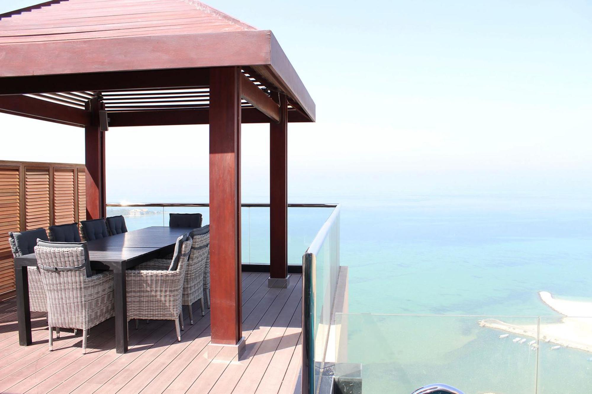 Lagoona Beach Luxury Resort And Spa Manama Ngoại thất bức ảnh The photo showcases a stylish outdoor patio or terrace area. It features a wooden structure with a sloped roof, providing a shaded space. There is a large table made of dark wood, surrounded by several wicker chairs. The patio is elevated and has gla