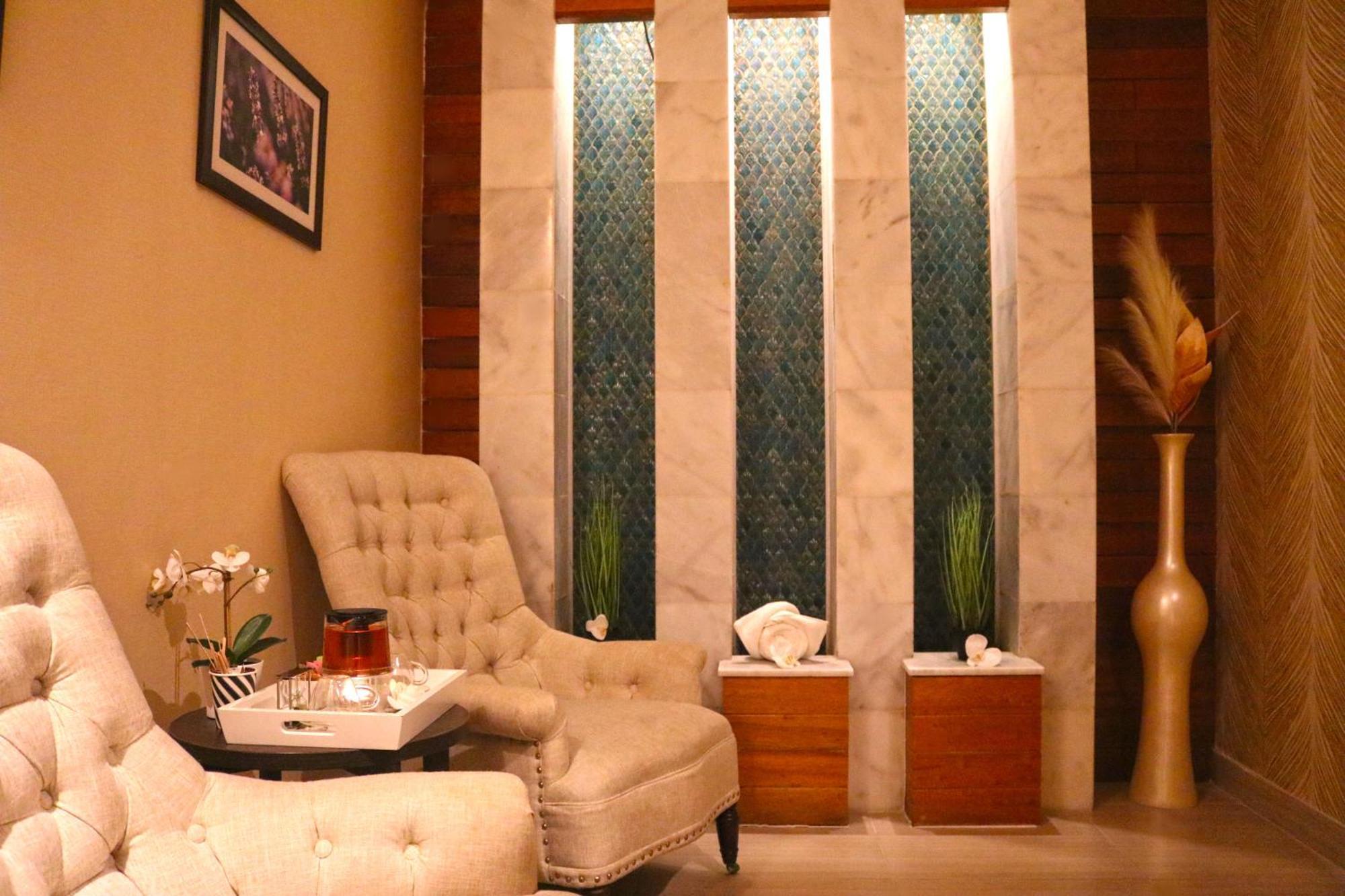 Lagoona Beach Luxury Resort And Spa Manama Ngoại thất bức ảnh The image shows a relaxing interior space, likely a waiting area or lounge. There are two plush, beige armchairs positioned near a small table, which holds a glass pitcher and a cup. The background features tall, elegant windows with decorative blue 