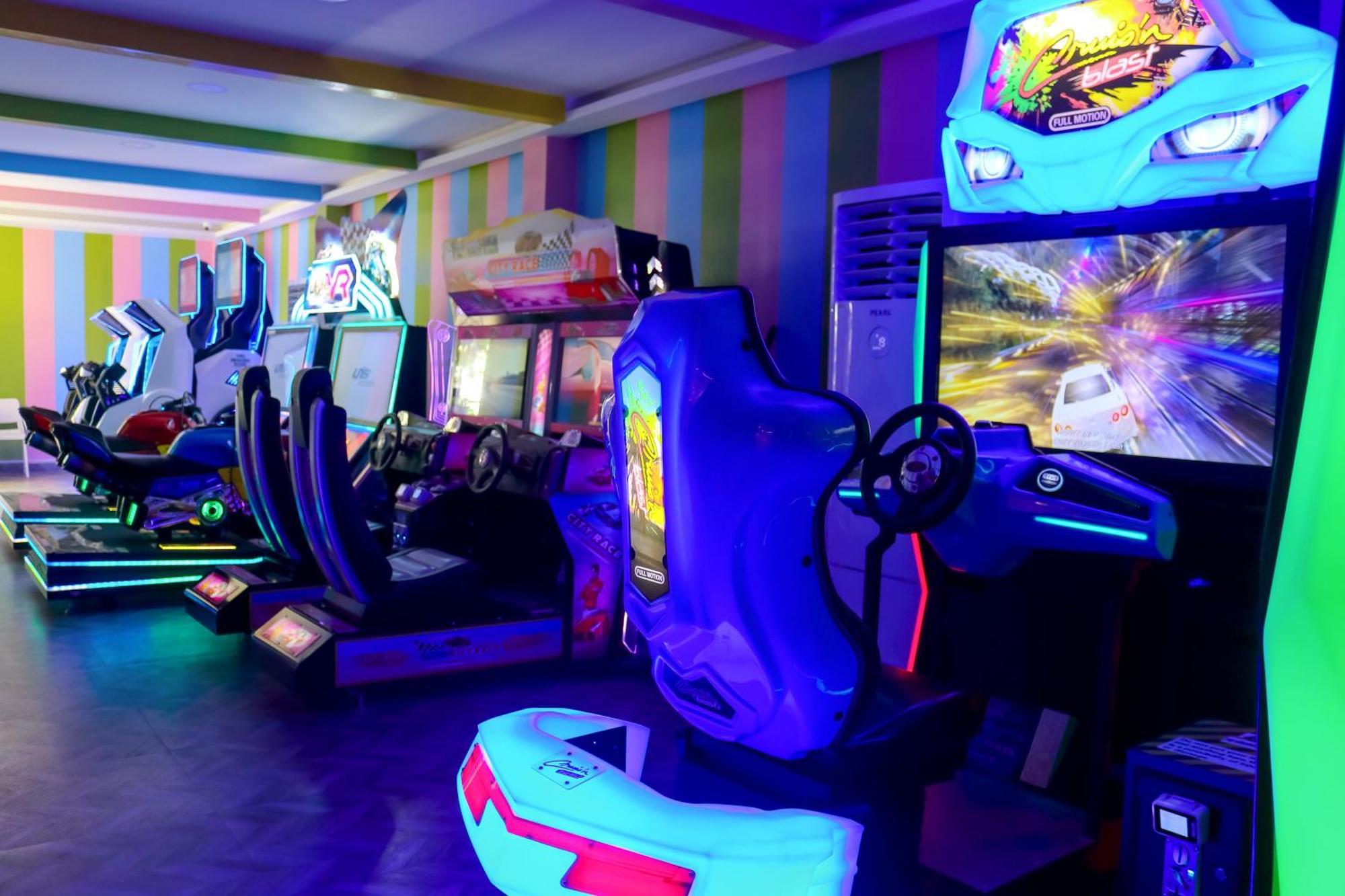 Lagoona Beach Luxury Resort And Spa Manama Ngoại thất bức ảnh The photo shows an arcade area filled with various arcade machines. The setting is illuminated with colorful neon lights, creating a vibrant atmosphere. There are several racing game machines with steering wheels and seats, each accompanied by screen