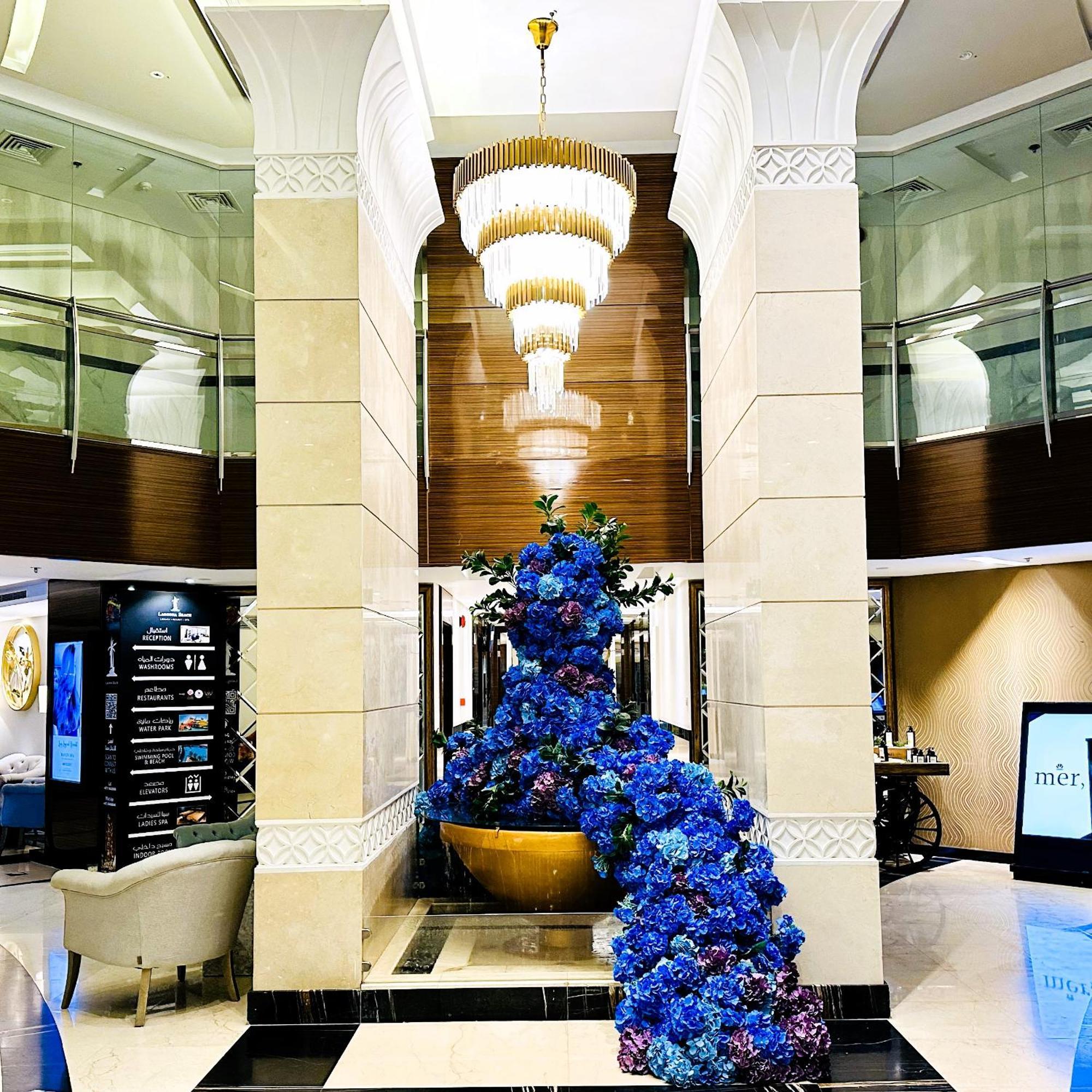 Lagoona Beach Luxury Resort And Spa Manama Ngoại thất bức ảnh The photo depicts an elegant lobby or entrance area of a building. Central to the scene is a large floral arrangement featuring vibrant blue flowers. This arrangement is positioned in a decorative bowl. Above it hangs a stylish chandelier with a laye