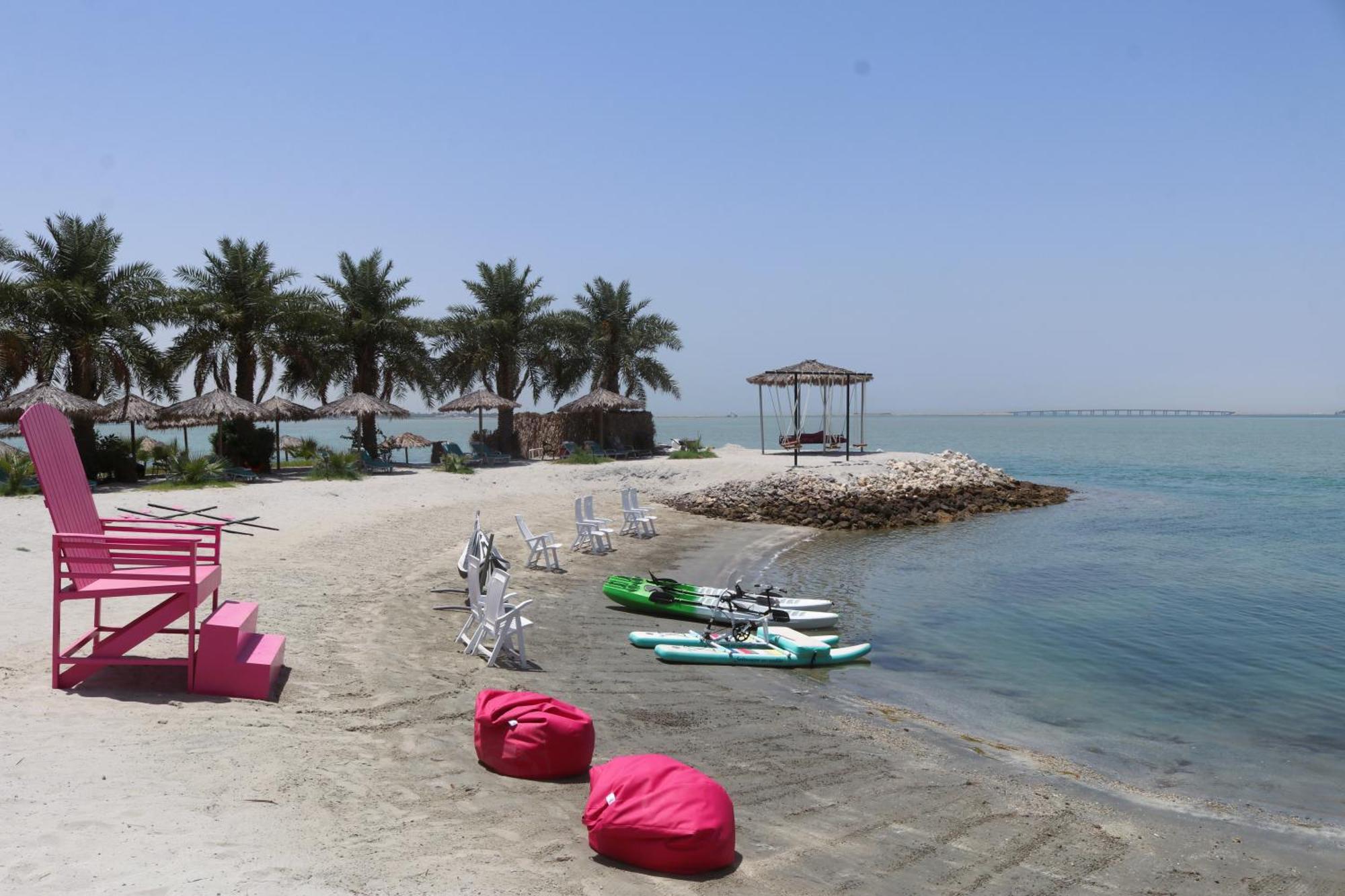 Lagoona Beach Luxury Resort And Spa Manama Ngoại thất bức ảnh The photo depicts a serene beach scene with clear blue water and a sunny sky. In the foreground, there are colorful pink bean bags and a large pink chair on the sand. A few white chairs and loungers are positioned near the water's edge. To the side, 