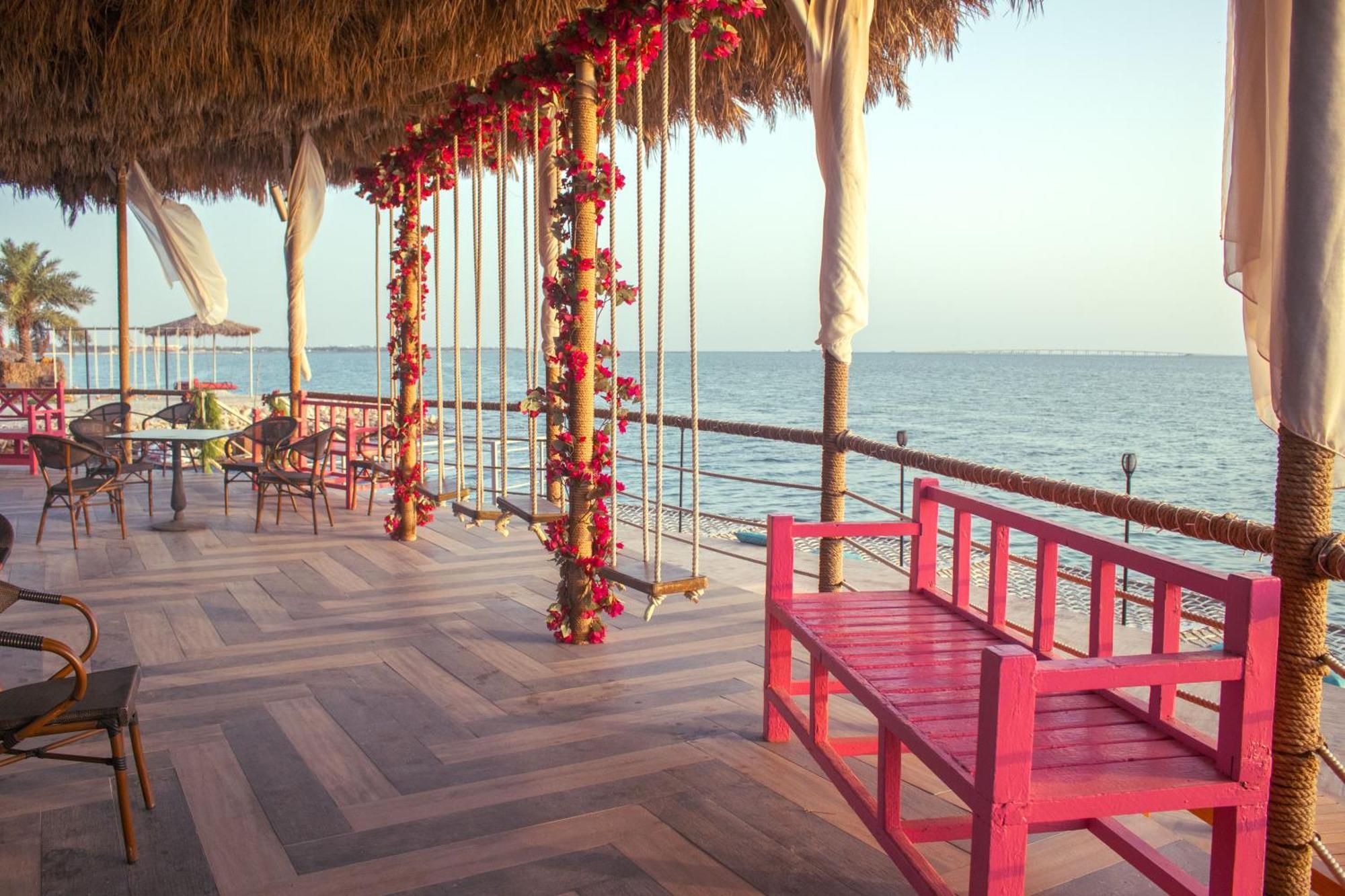 Lagoona Beach Luxury Resort And Spa Manama Ngoại thất bức ảnh The photo depicts a scenic outdoor area by the water, with a wooden deck and a thatched roof. Two swings adorned with flowers are hanging from the ceiling, providing a charming touch to the space. In the foreground, there is a bright pink bench, addi
