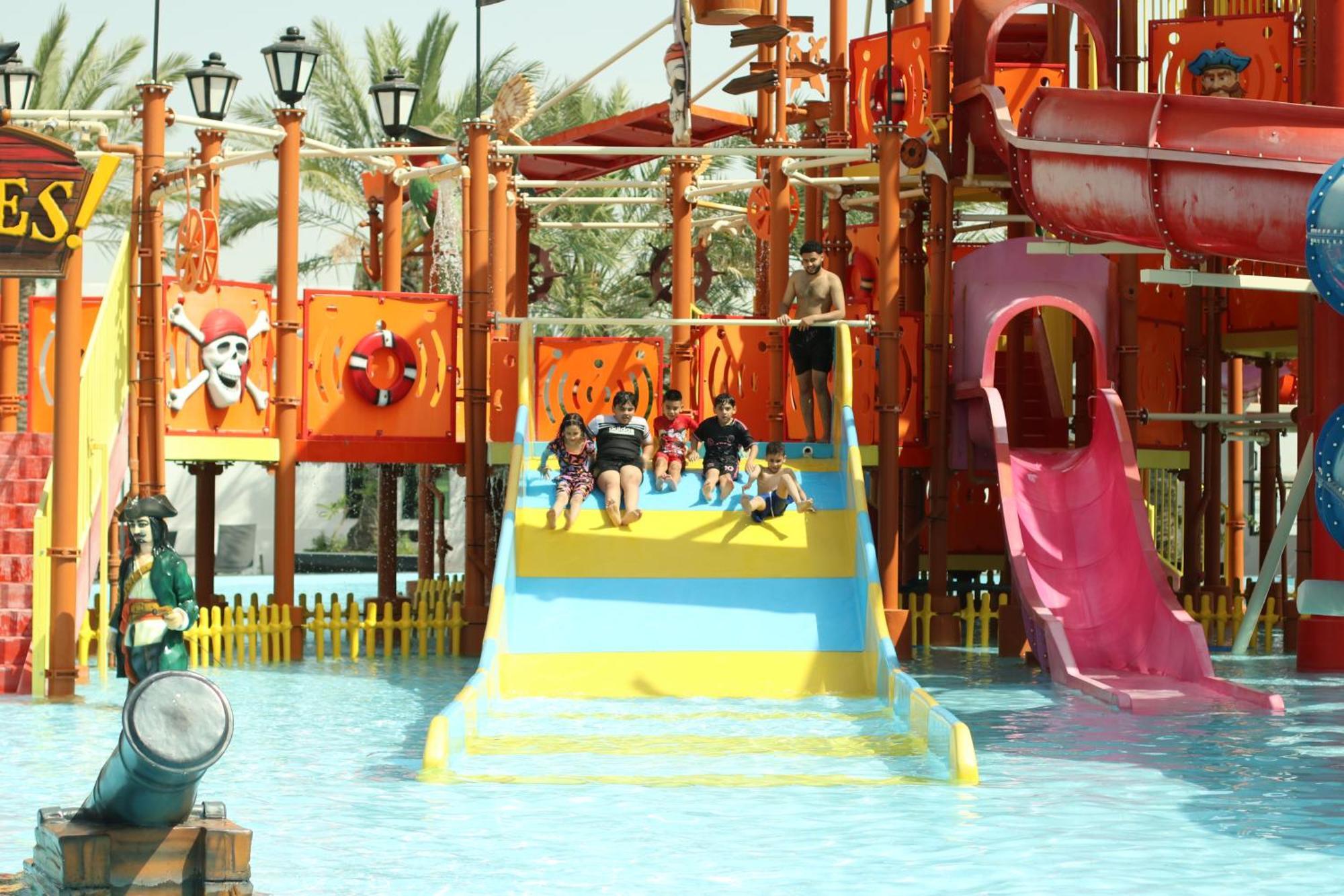 Lagoona Beach Luxury Resort And Spa Manama Ngoại thất bức ảnh The photo shows a colorful water park scene featuring a play area with multiple slides. There are children enjoying themselves, sliding down bright yellow and pink slides into a pool of water. The structure has pirate-themed decorations, including a 