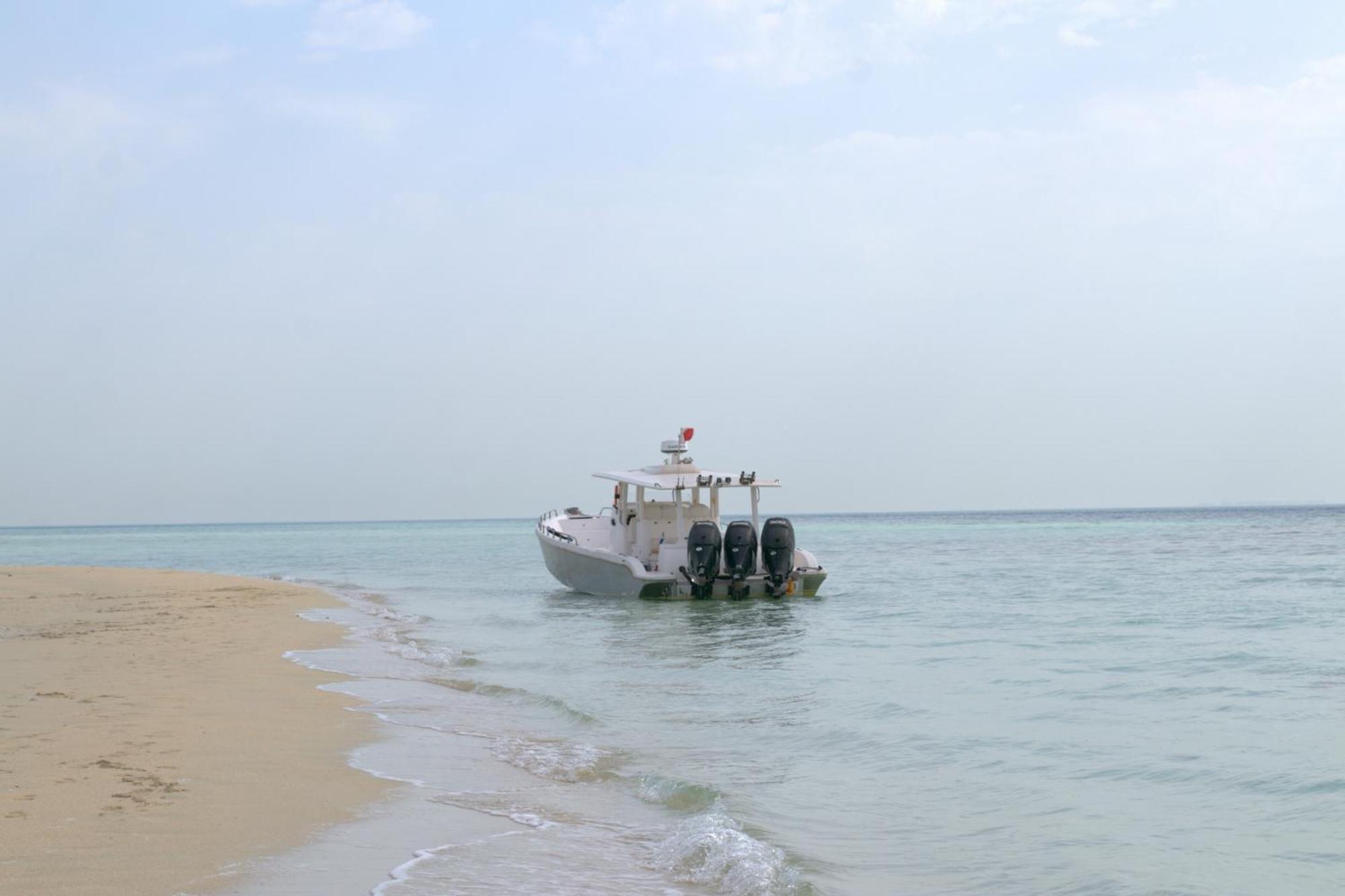 Lagoona Beach Luxury Resort And Spa Manama Ngoại thất bức ảnh The photo depicts a small boat anchored near a sandy beach, partially submerged in calm, clear water. The boat is white and features three outboard motors on its stern. In the background, the sea stretches out toward the horizon under a light, cloudy
