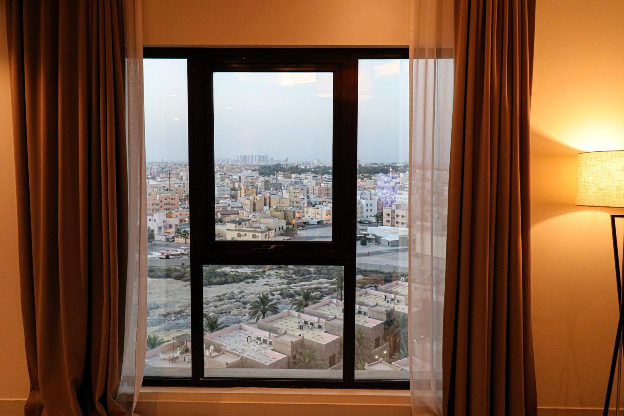 Lagoona Beach Luxury Resort And Spa Manama Ngoại thất bức ảnh The photo shows a window with a black frame and sheer curtains. Through the window, there is a view of a cityscape, displaying a densely populated area with various buildings and rooftops. The sky appears slightly overcast, suggesting a muted lightin