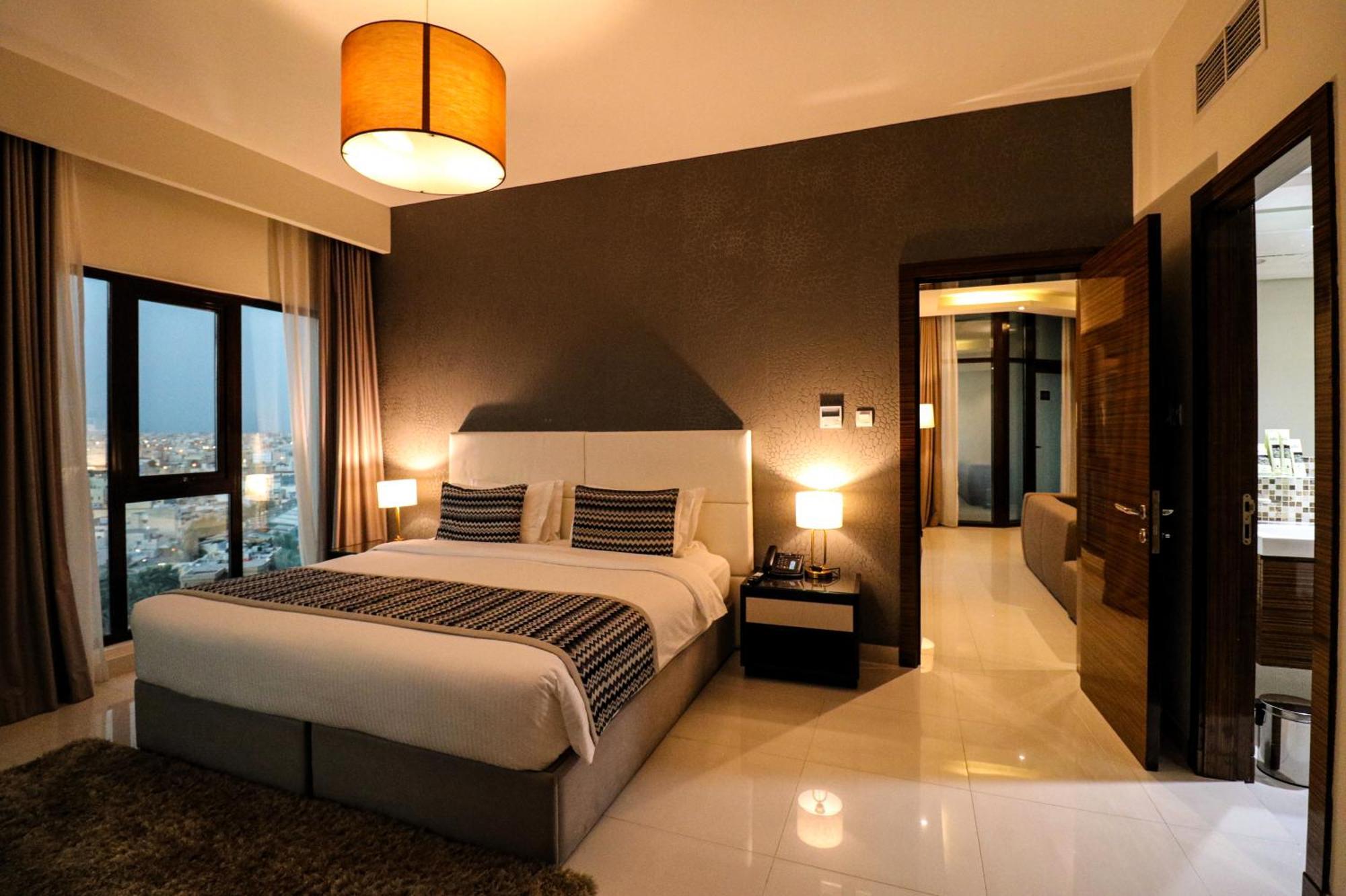 Lagoona Beach Luxury Resort And Spa Manama Ngoại thất bức ảnh The photo depicts a modern hotel room or suite. It features a large bed with a white and gray color scheme, accented with decorative pillows. On either side of the bed, there are sleek bedside lamps providing warm lighting. The walls are a dark color