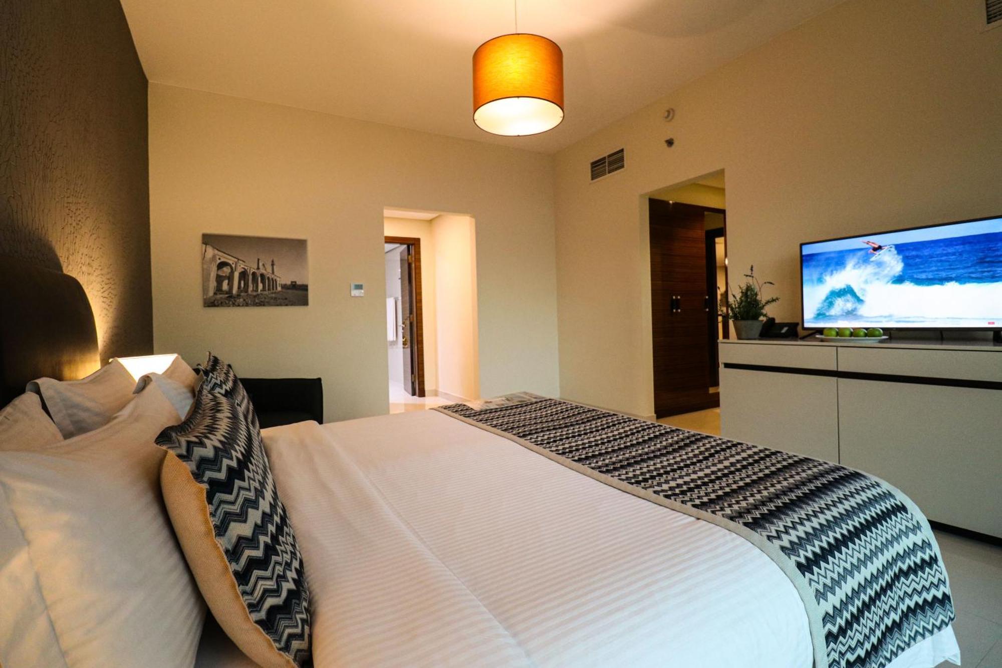 Lagoona Beach Luxury Resort And Spa Manama Ngoại thất bức ảnh The photo shows a well-decorated hotel room. There is a large bed with white linens and decorative pillows featuring a zigzag pattern. On the wall beside the bed is a piece of artwork, and there is a modern television on a white furniture unit opposi