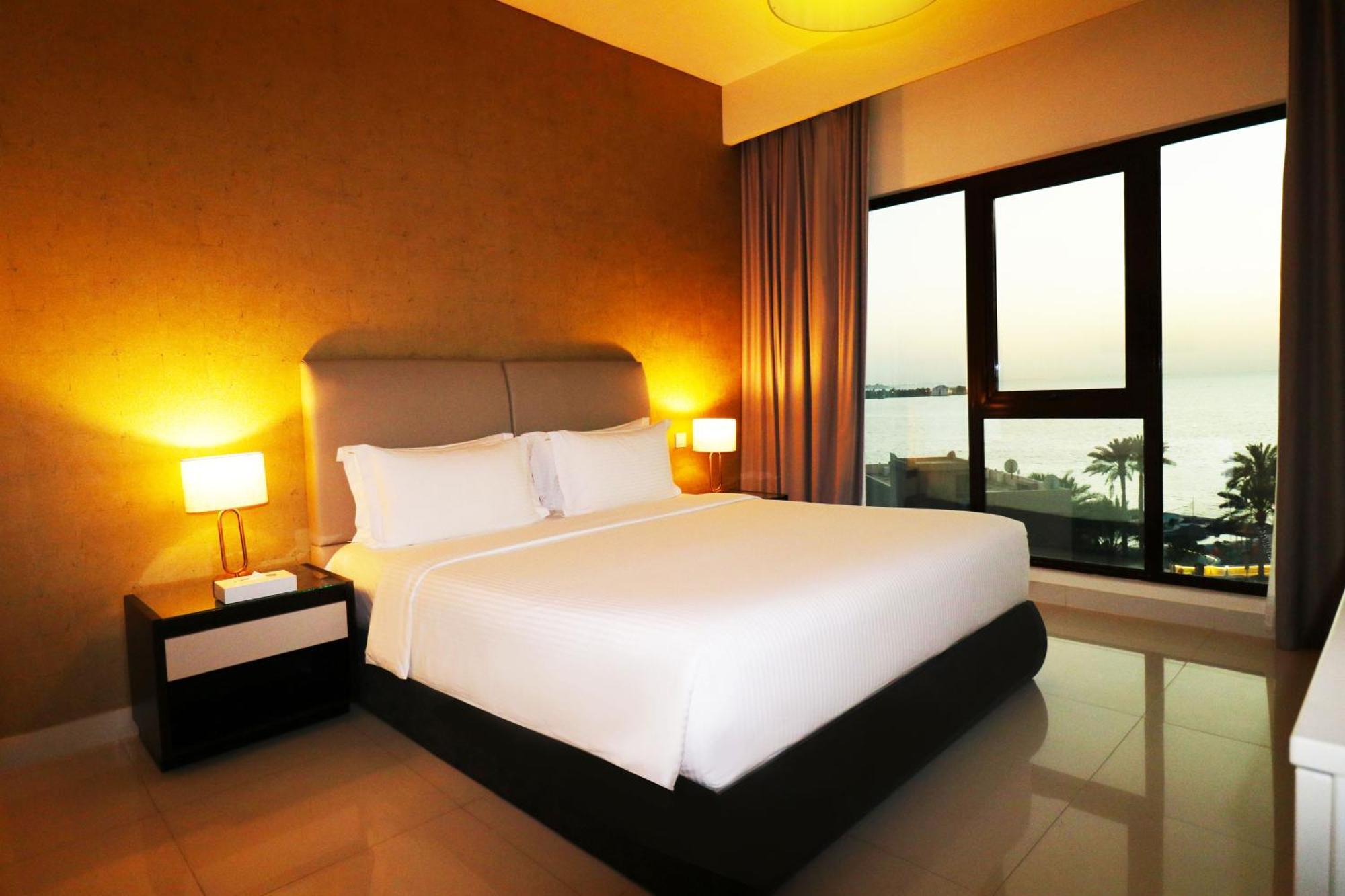 Lagoona Beach Luxury Resort And Spa Manama Ngoại thất bức ảnh The photo shows a modern hotel bedroom with a large, comfortable bed dressed in white linens. The room features warm lighting, including bedside lamps, creating a cozy atmosphere. There are large windows with a view of the outdoors, possibly overlook
